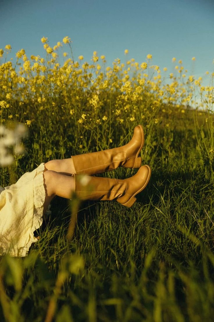 A person lying on a grassy field with brown boots, wearing a cream-colored dress, against a backdrop of yellow flowers and a clear blue sky.
