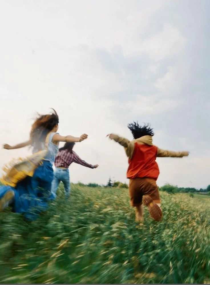 Four kids running through a grassy field on a cloudy day.