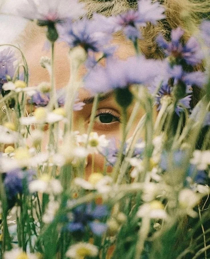 A person with blonde hair peering through a dense arrangement of purple and white flowers.