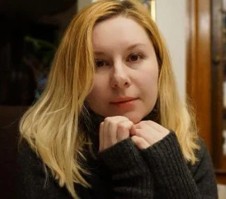 A young woman with light skin and blonde hair wearing a dark sweater, resting her chin on her hands, in an indoor setting.