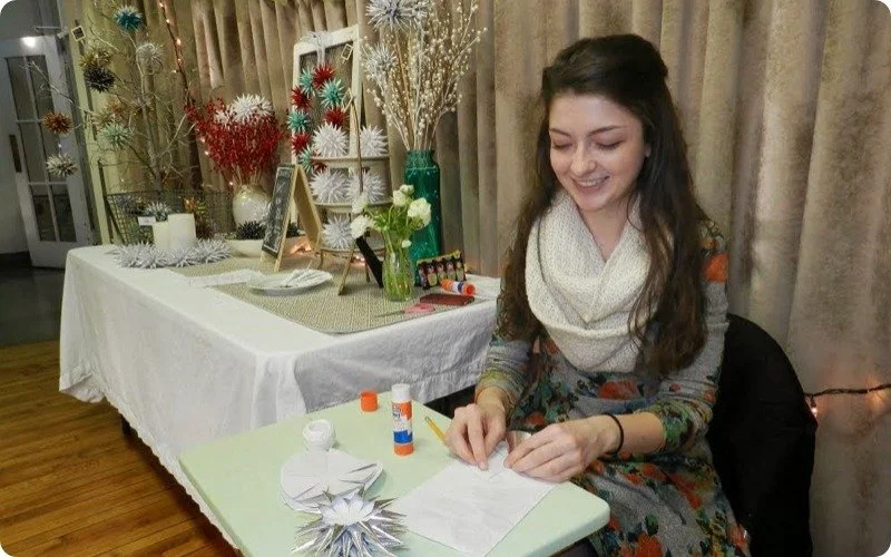 A young woman with long dark hair, wearing a patterned dress and white scarf, is sitting at a table with holiday crafts, smiling while working on a paper project. Behind her is a decorated table with Christmas ornaments, white flowers, greenery, and festive decorations, and curtains in the background.