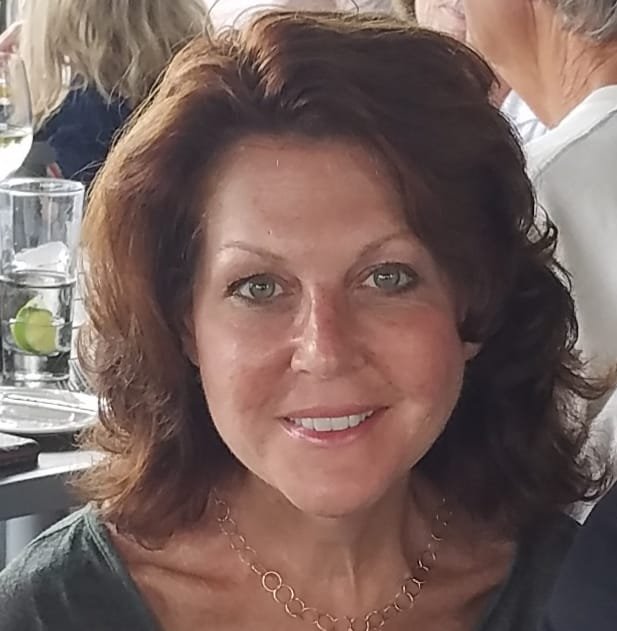 A woman with shoulder-length brown hair smiling at a social event, with glasses of water and lime slices on the table behind her.