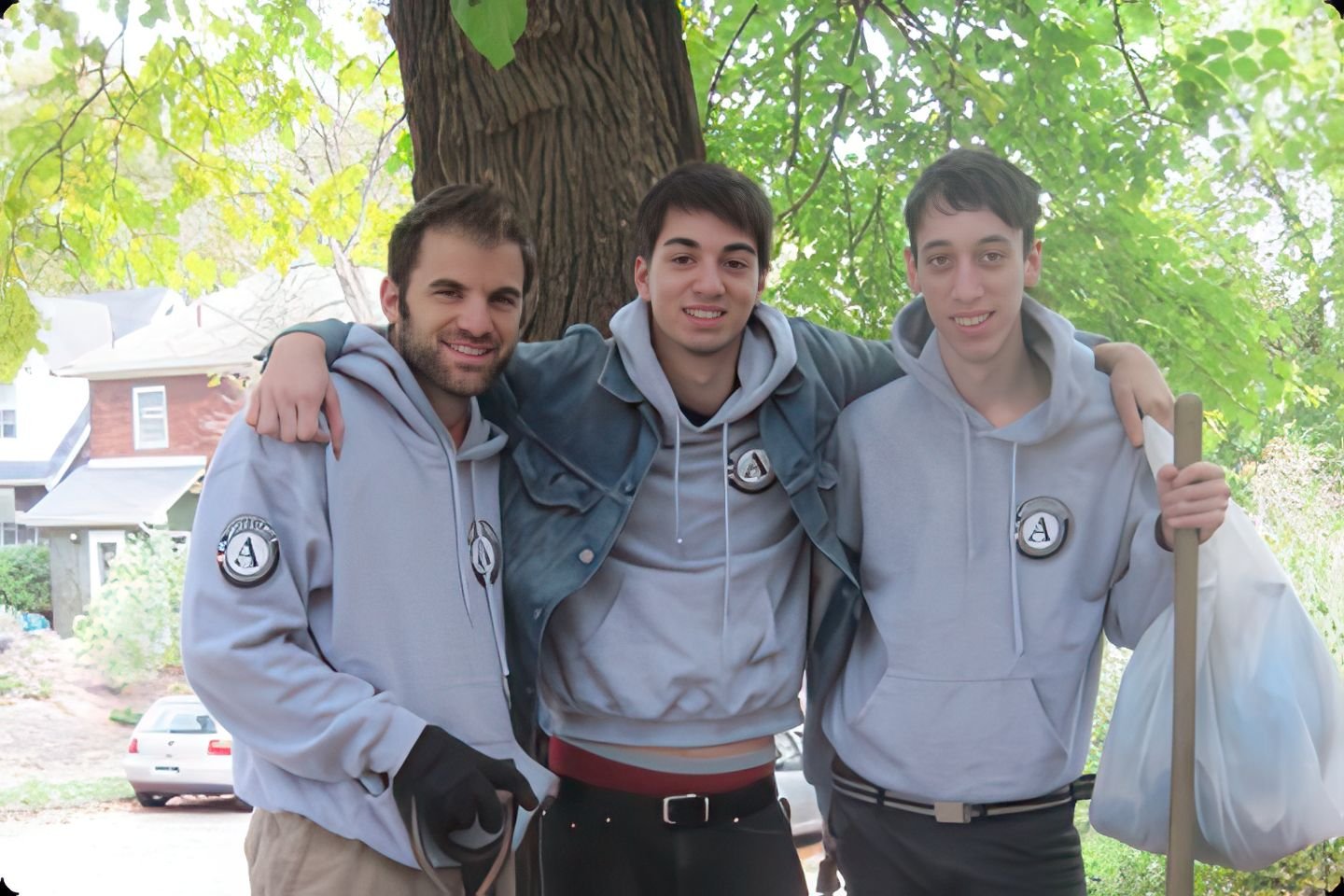 Three young men standing together outdoors under a large tree, smiling, wearing matching gray hoodies with a logo on the chest, with one holding a broom and a bag.