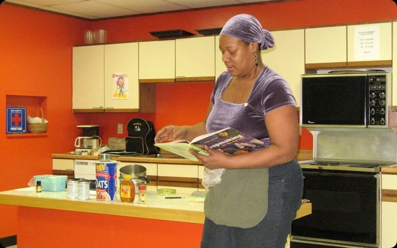 Woman wearing a purple shirt and headscarf reading a cookbook in a kitchen with orange walls and cream cabinets.