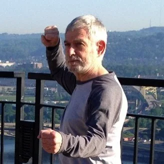 Man with short gray hair and beard making a fighting pose outdoors on a rooftop with a cityscape in the background.