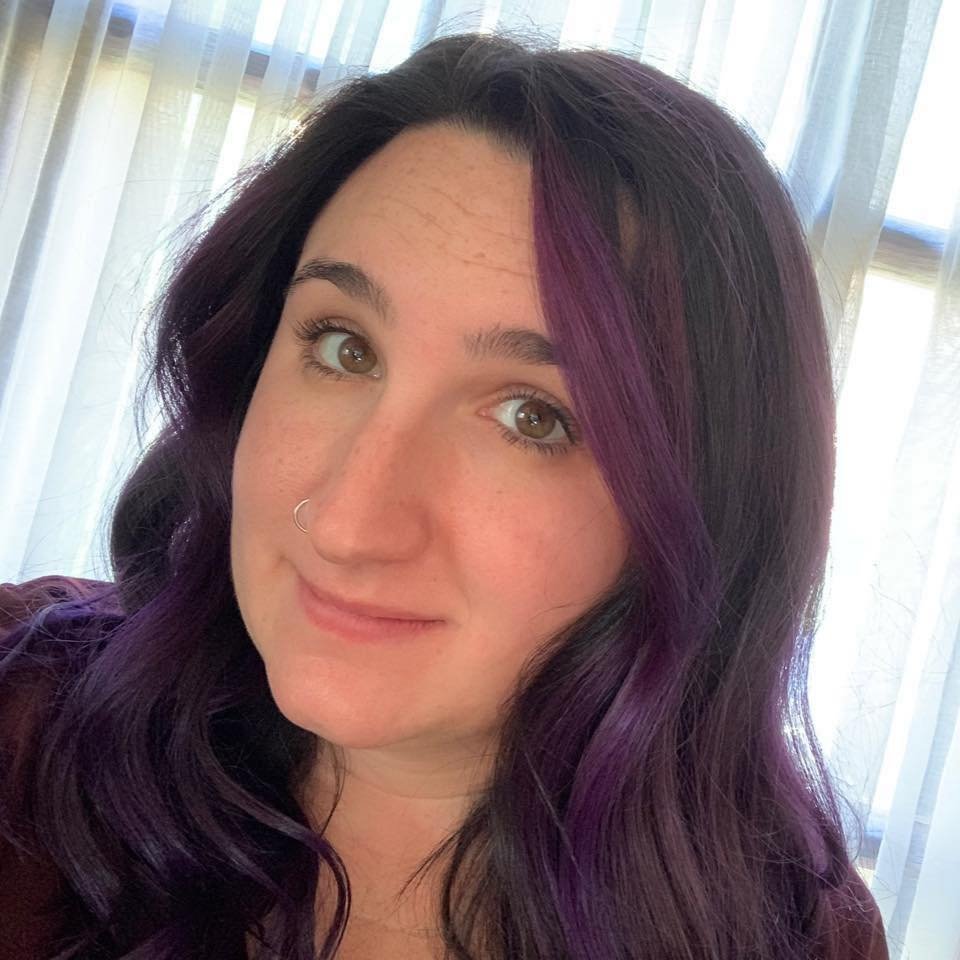 A woman with purple highlights in her dark hair, light skin, and hazel eyes, smiling gently, in front of light-colored curtains.