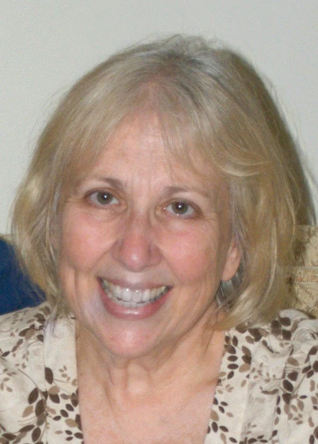 A smiling middle-aged woman with blonde hair, wearing a beige top with brown leaf patterns, sitting indoors near a white wall.
