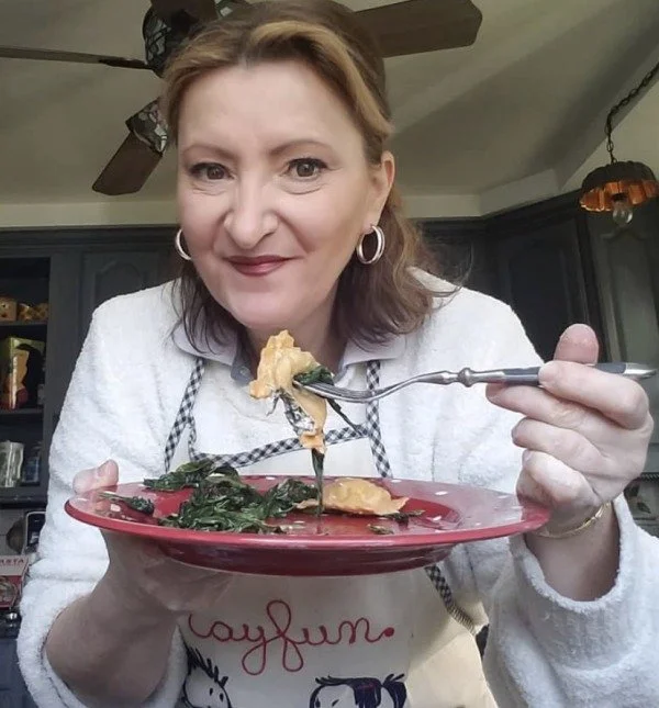 A woman holding a red plate with cooked greens and a piece of pie, smiling at the camera in a kitchen.