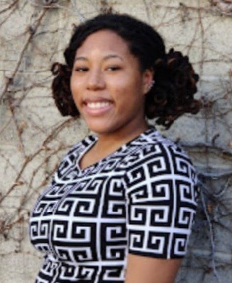 A smiling woman with dark curly hair posing outdoors in front of a wall with vines, wearing a patterned black and white top.