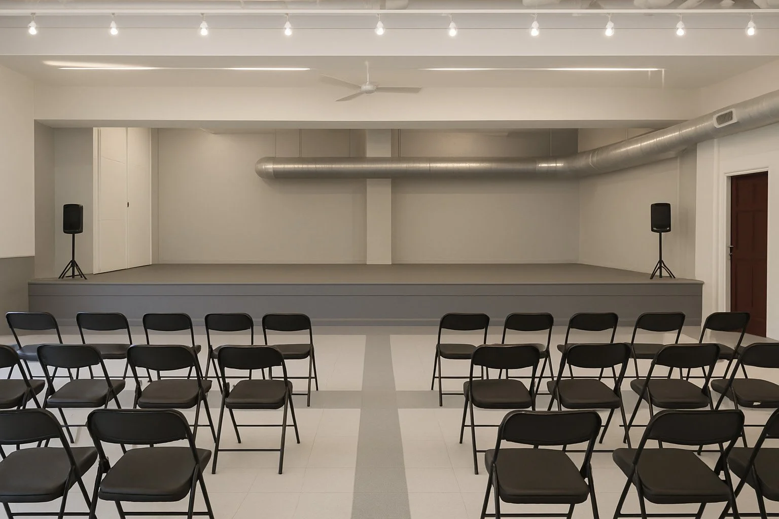 Empty indoor stage with two black speakers, a ceiling fan, string lights, and rows of black chairs facing the stage.