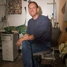 A man with a beard and short hair sitting on a black office chair in a cluttered room, smiling at the camera.