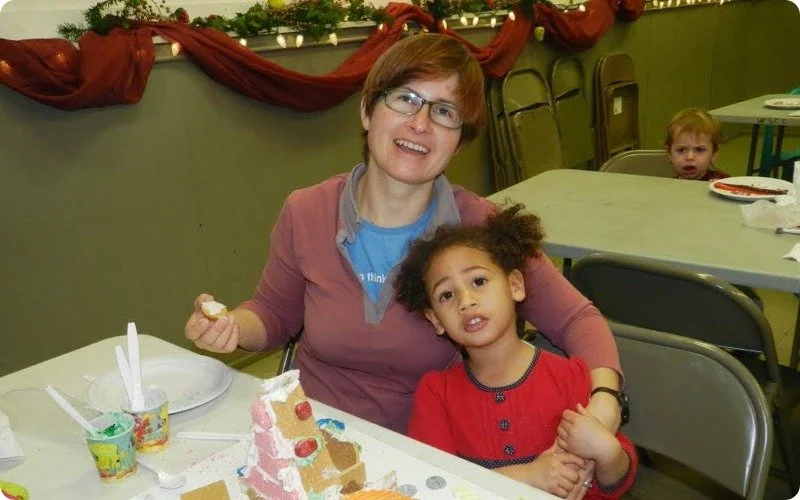 A woman with glasses and short hair, smiling, sits with a young girl with curly hair, at a decorated table with a gingerbread house, during a celebration or party. There is a little boy with red hair in the background at another table.