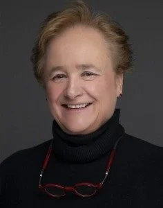 Portrait of an older woman with short red hair, smiling, wearing a black turtleneck and glasses hanging around her neck, against a dark background.