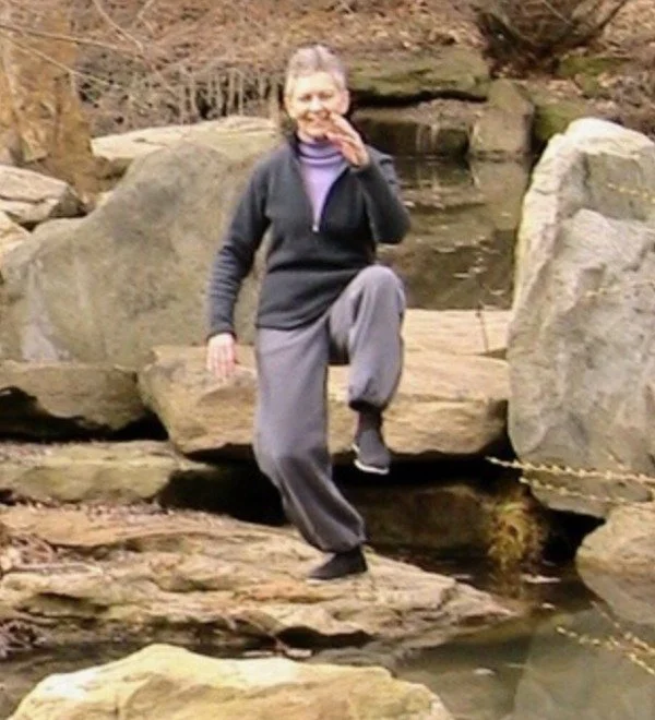 A woman balancing on rocks near a creek, with one foot on a rock and her hand covering her mouth.