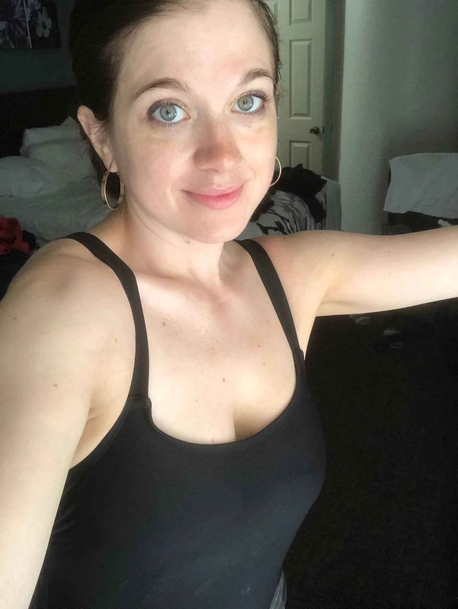 A woman with light skin, green eyes, and dark hair taking a selfie in a room with beds and a door visible in the background. She is wearing a black tank top and hoop earrings.