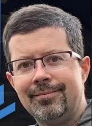 Close-up of a man with short dark hair, glasses, and facial hair, smiling at the camera.