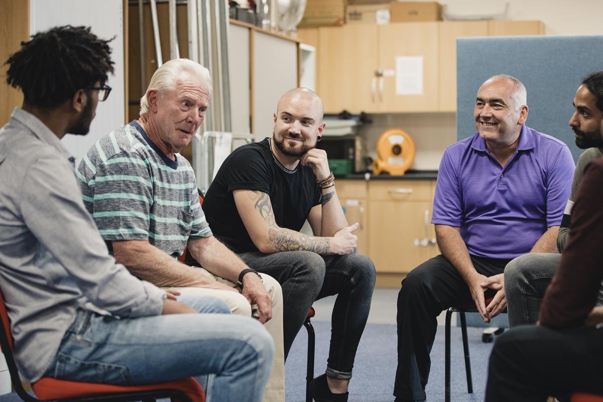 A diverse group of six men sitting in a circle in a casual setting, engaged in a conversation, with a kitchen area in the background.
