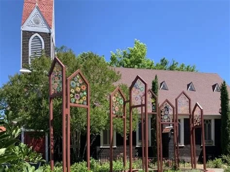 Photo of some public art backed with the side of a church. The art looks like rusty metal shapes, about 12 feet tall, echoing the shape of the church's tower. Colorful shapes are suspended within the metal frames.
