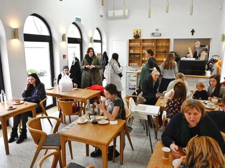 A busy café with people sitting at wooden tables, some eating, some drinking, and children with adults. There are large arched windows on the left, and staff behind the counter in the back area.