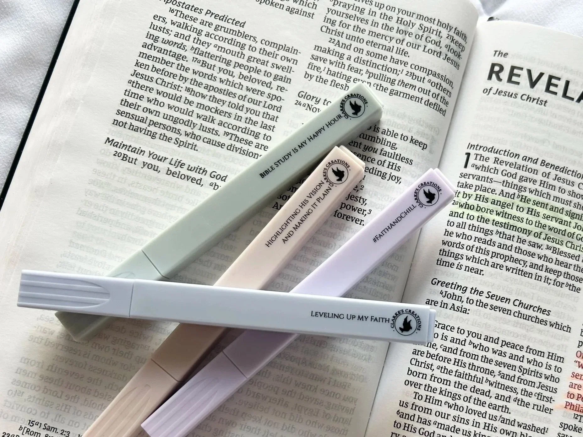 Open Bible with four highlighters resting on it. The Bible is open to a page about the Revelation of Jesus Christ. The highlighters are pastel colors with the brand name 'Chalkos Creations' and phrases like 'Bible Study is my Happy Hour,' 'Highlighting His Vision and Making It Plain,' '#FaithAndChill,' and 'Leveling Up My Faith' written on them.