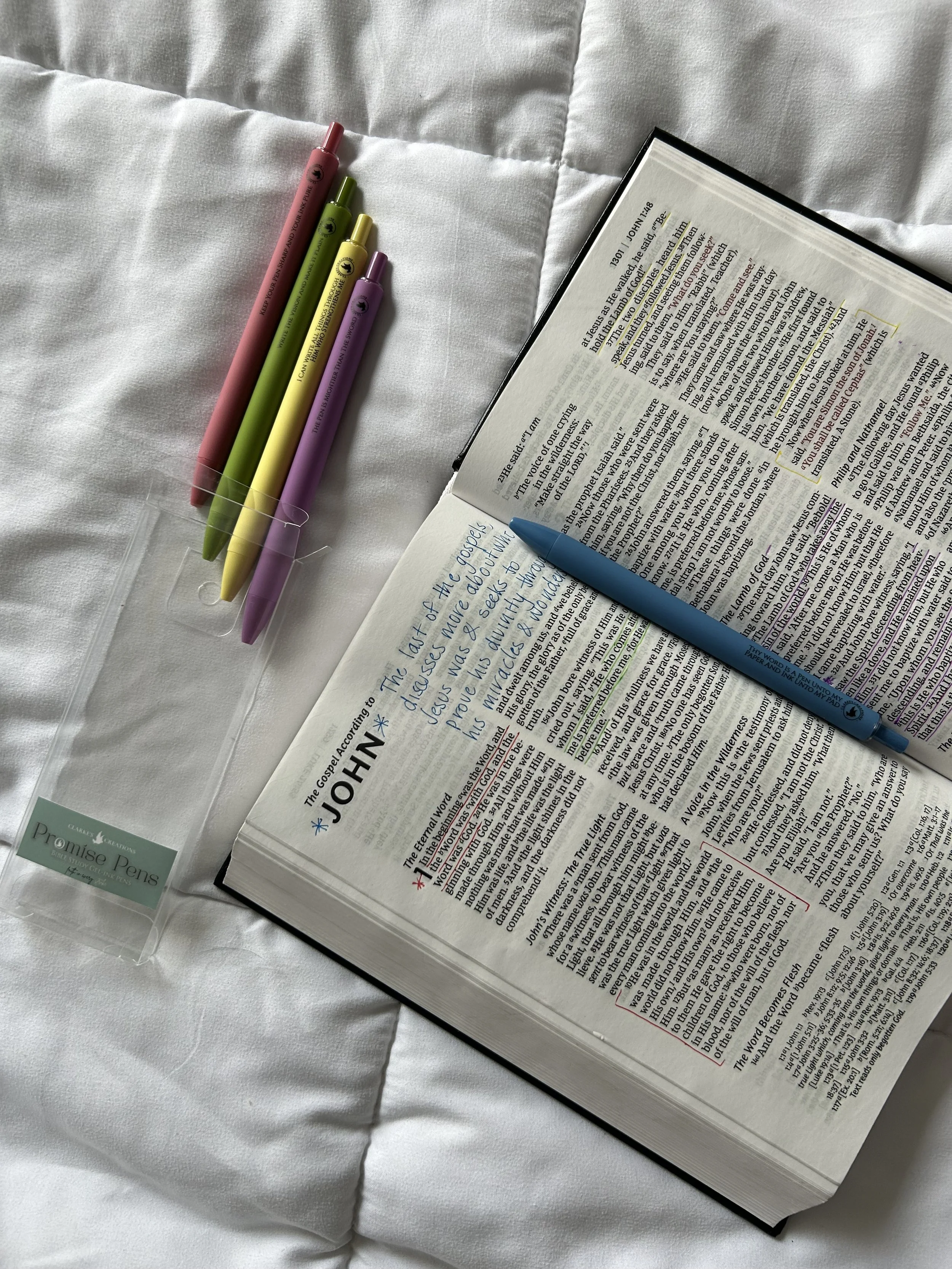 An open Bible with colorful pens, with Christian phrases inscribed on the shafts. Handwritten notes using the pens are on the pages about John in the New Testament, placed on a white bedspread.