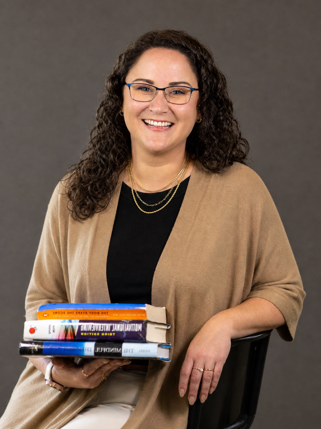 A female licensed professional counselor is pictured with a warm smile wearing a black top, gold jewelry, and holding three books: "The mindful Therapist", "Motivational Interviewing" and "The body keeps the Score".
