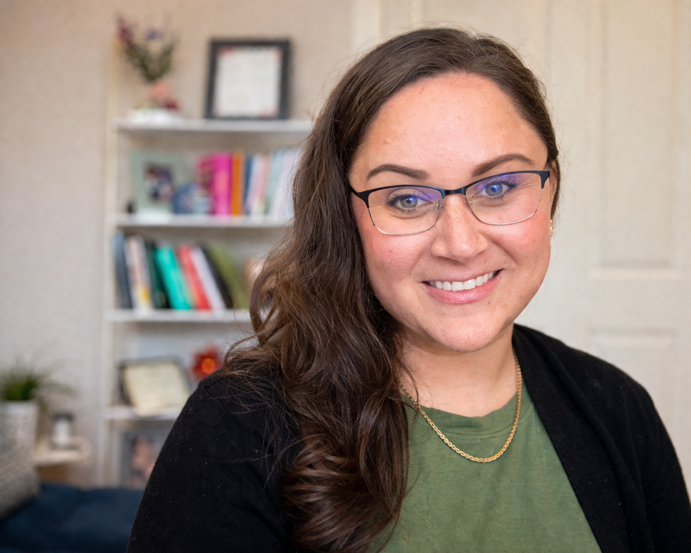 A smiling woman with glasses, long wavy brown hair, wearing a green shirt, black cardigan, and a gold necklace in a room with bookshelf and framed picture in the background.