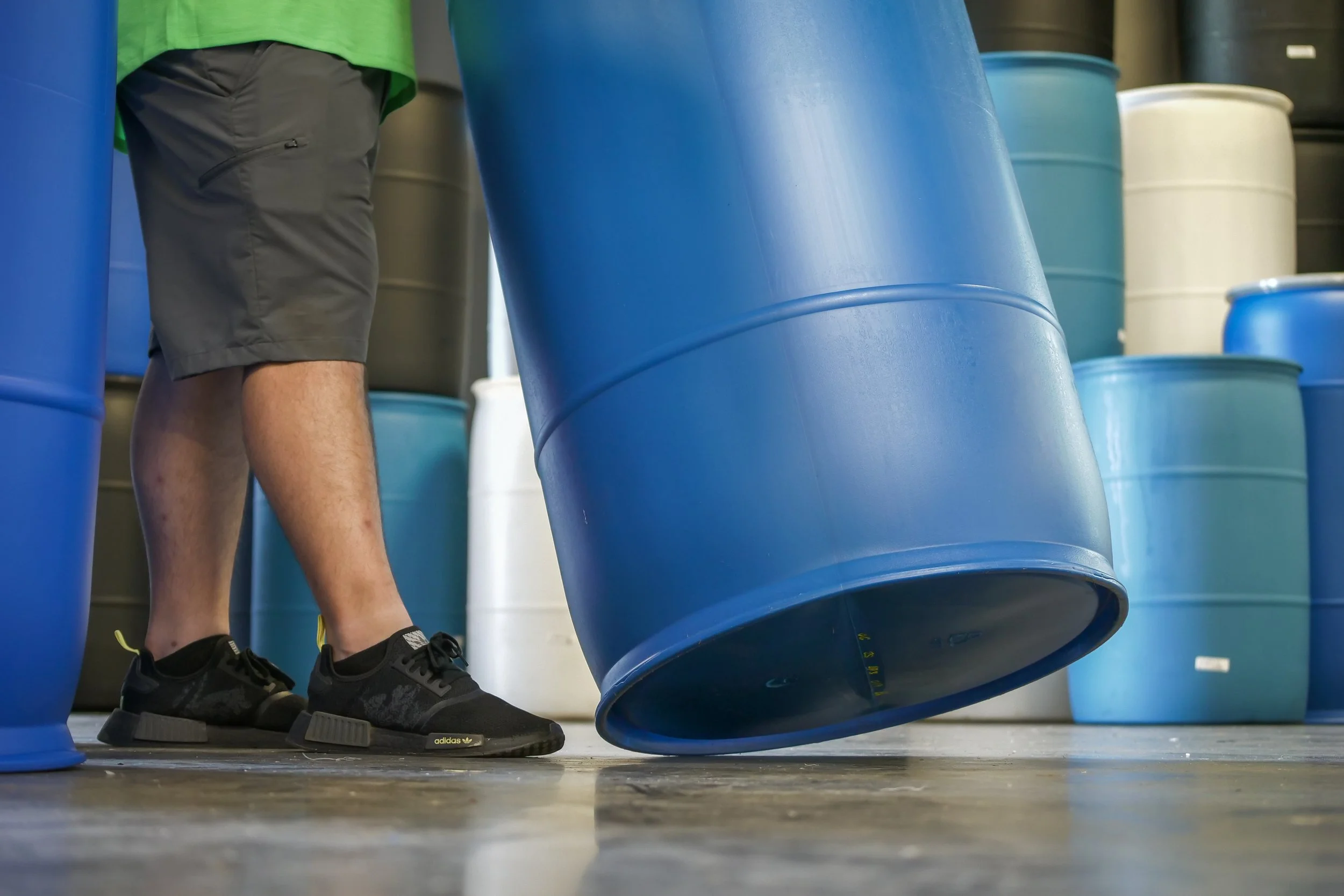 A person wearing black sneakers and gray shorts is lifting a large blue plastic barrel in a storage room with other barrels in the background.