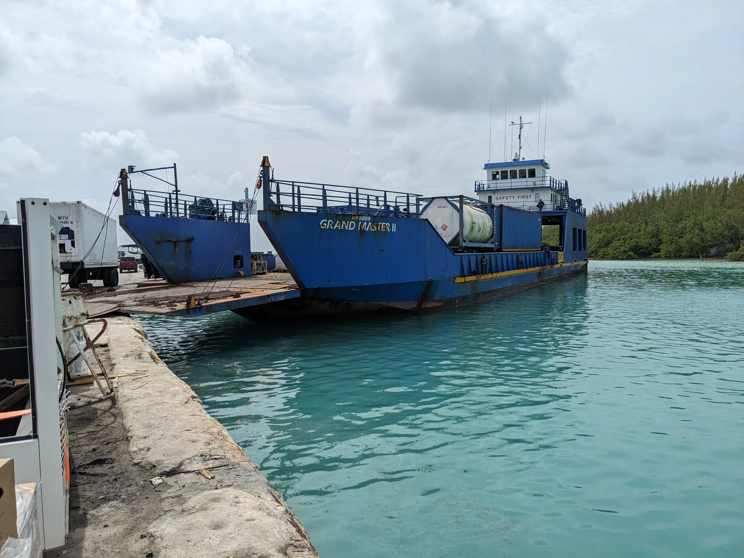 Blue ferry boat named Grand Master II docked at a pier with calm turquoise water, cloudy sky, and green trees in the background