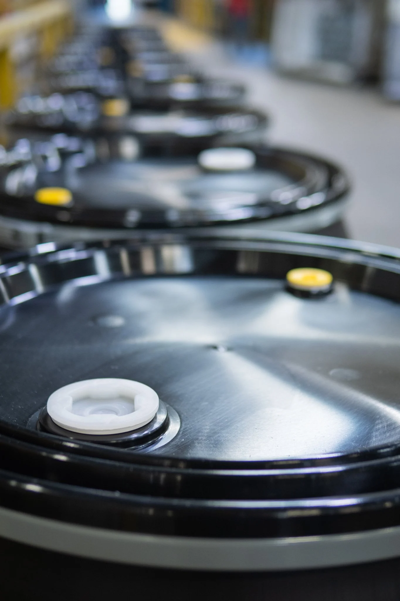 A row of black industrial plastic drums with white and yellow lids positioned on a factory floor.