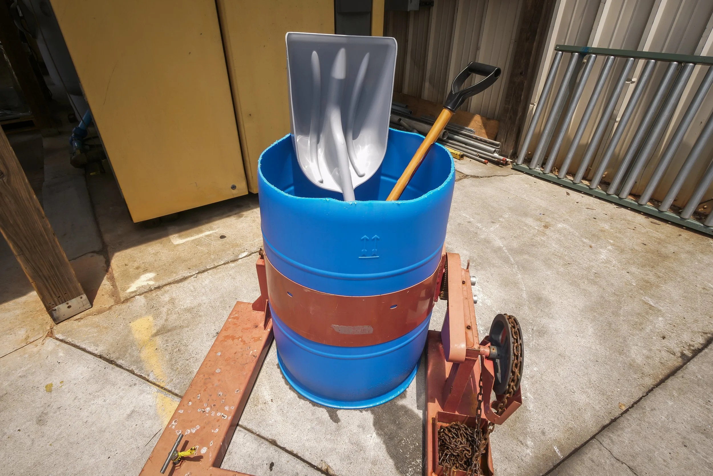 A blue plastic barrel with a metal scoop and a gardening shovel inside, mounted on a moving dolly with a chain and gears, outdoors on a concrete surface.