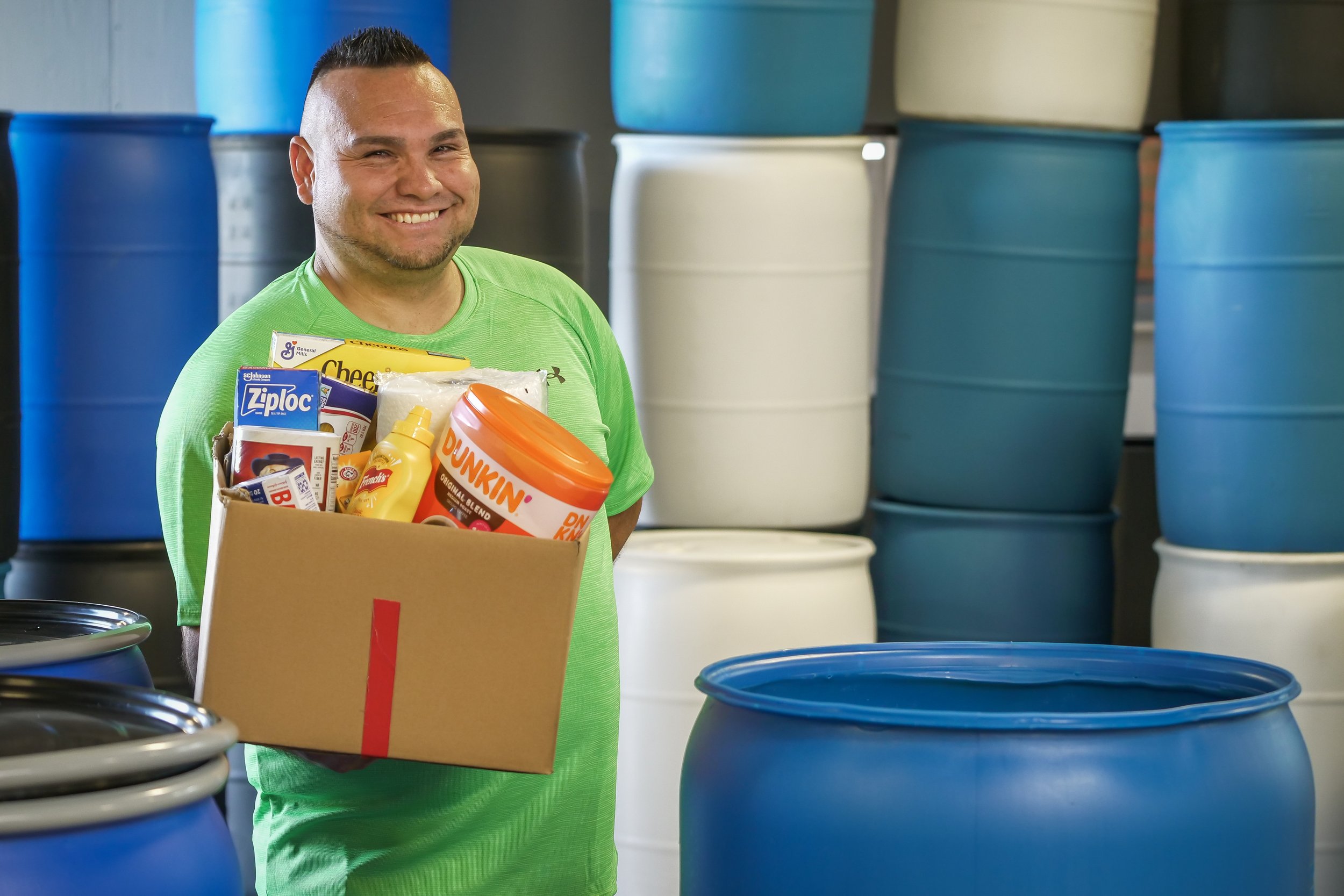 A smiling man in a green shirt holding a cardboard box filled with food items, standing in front of large blue and white barrels in a storage or warehouse setting.