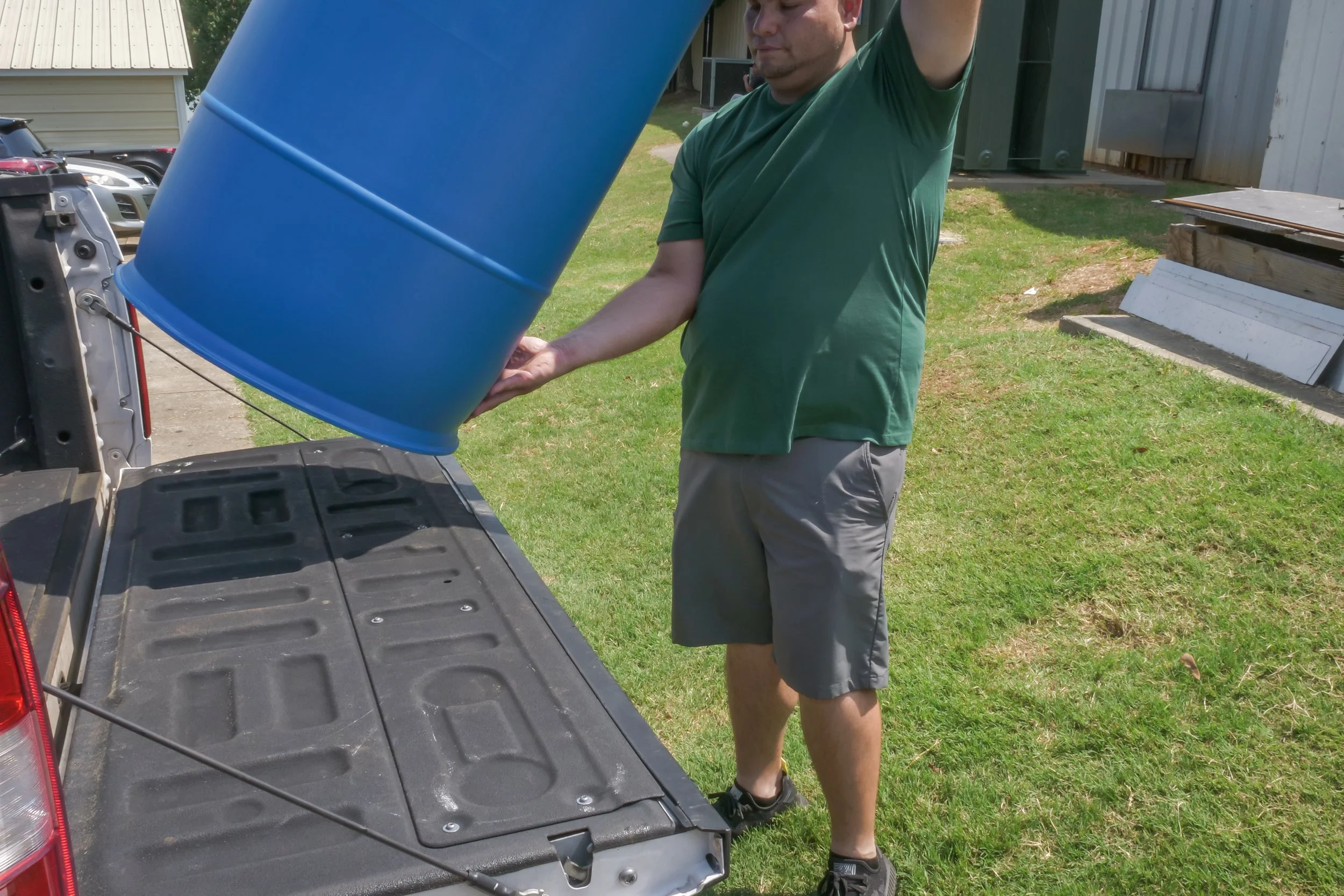 A man in a green shirt and gray shorts pouring a blue barrel into the back of a pickup truck in a grassy area.