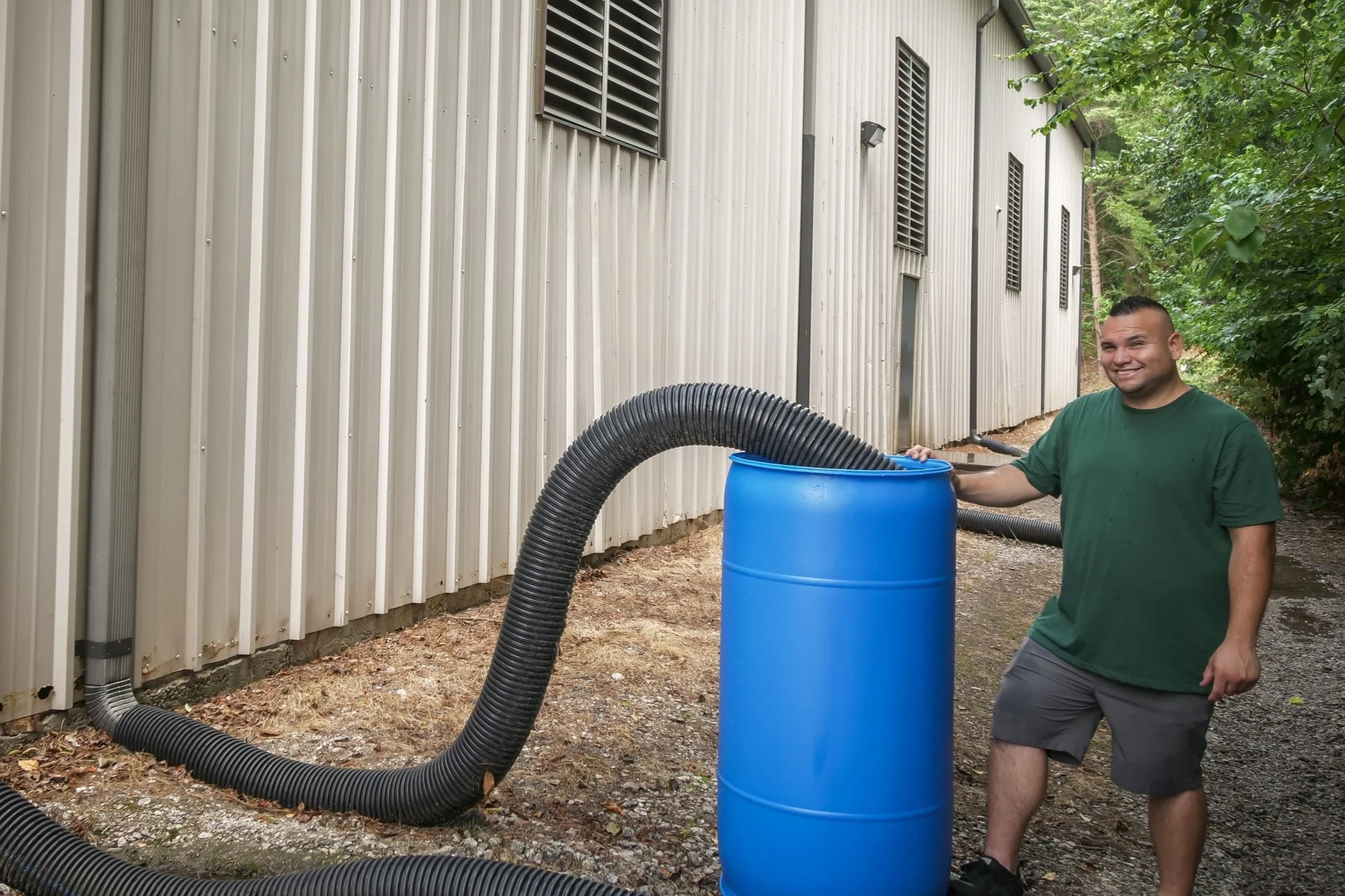 A man standing outdoors near a metal building with a large blue barrel and black flexible hoses connected to it, smiling at the camera.
