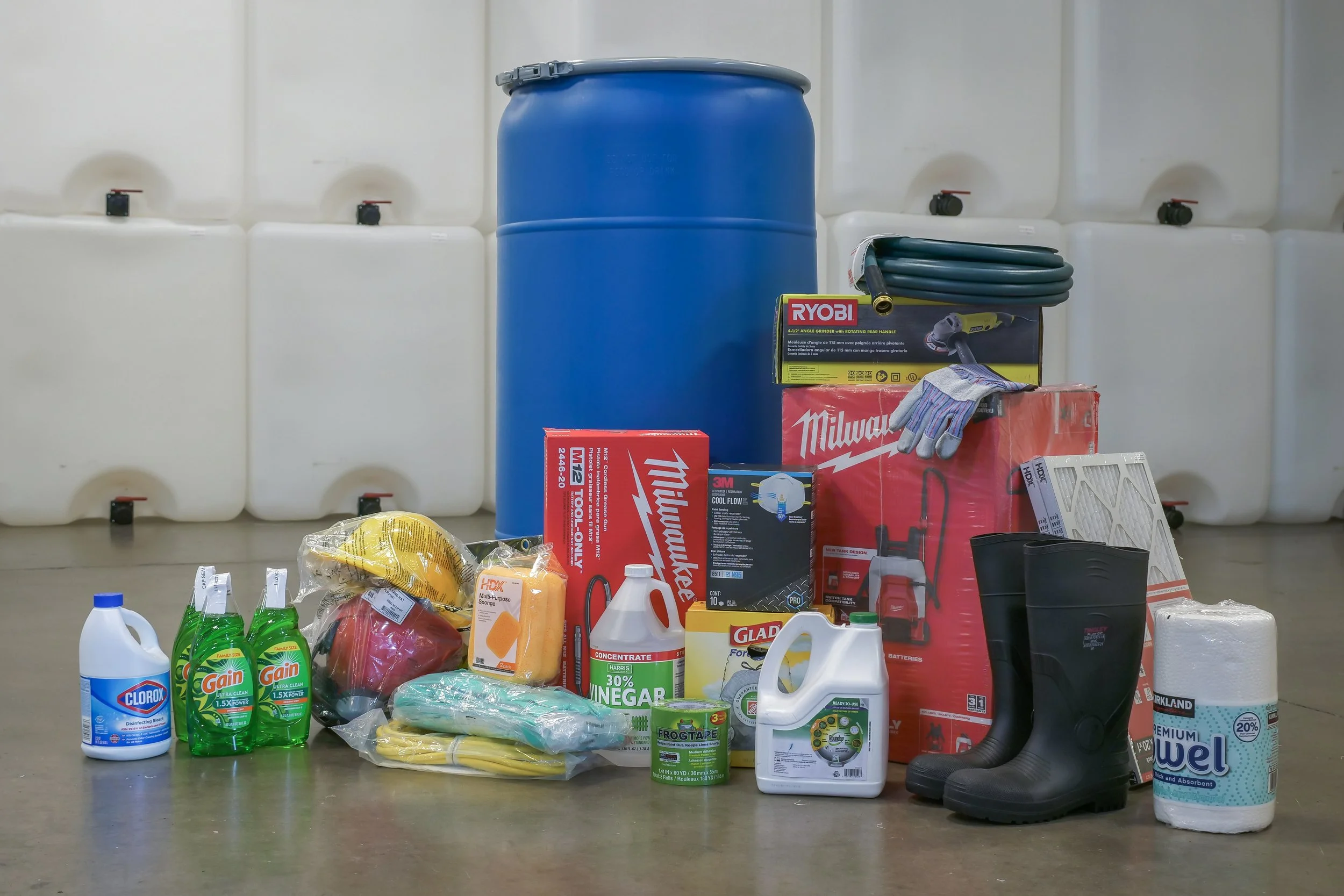 Cleaning supplies and tools, including bleach, dish soap, gloves, boots, and a large blue barrel, arranged on the floor in front of white containers.