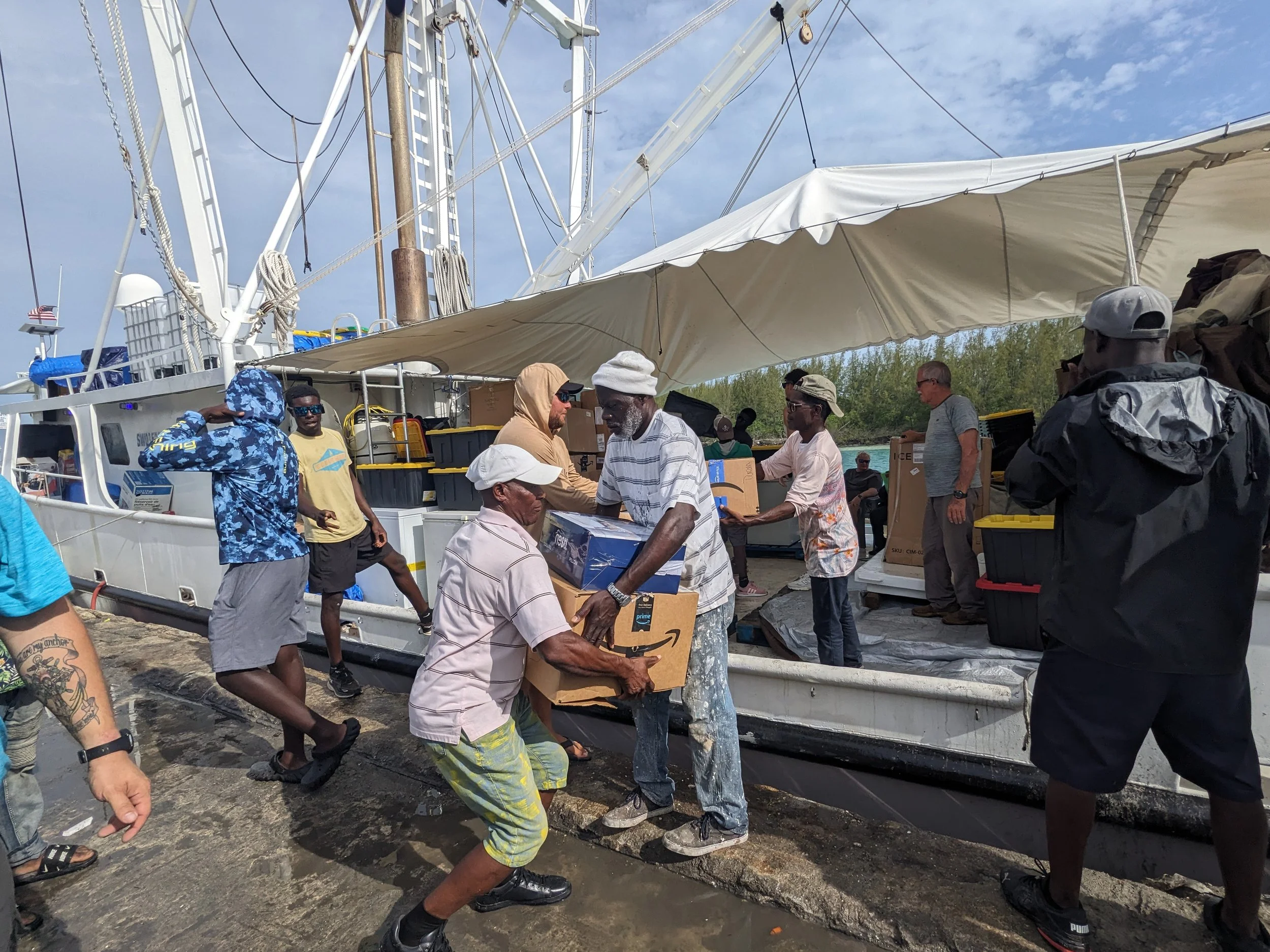Group of people loading supplies onto a boat docked at a pier, with some individuals handing over boxes and bags, under a blue sky with white clouds.