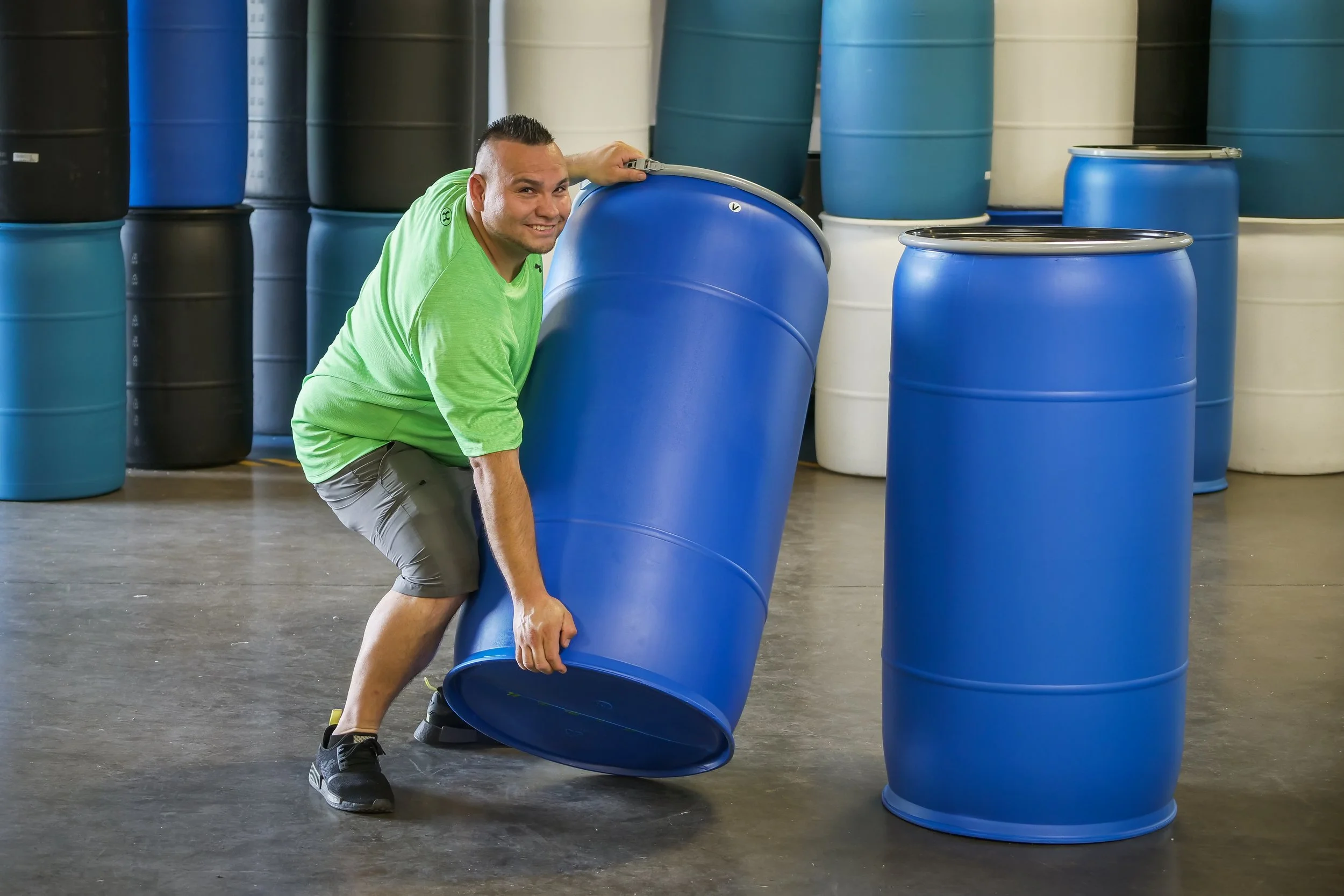 A man wearing a green shirt and shorts is lifting a large blue barrel in a warehouse. There are many similar barrels behind him in various colors.