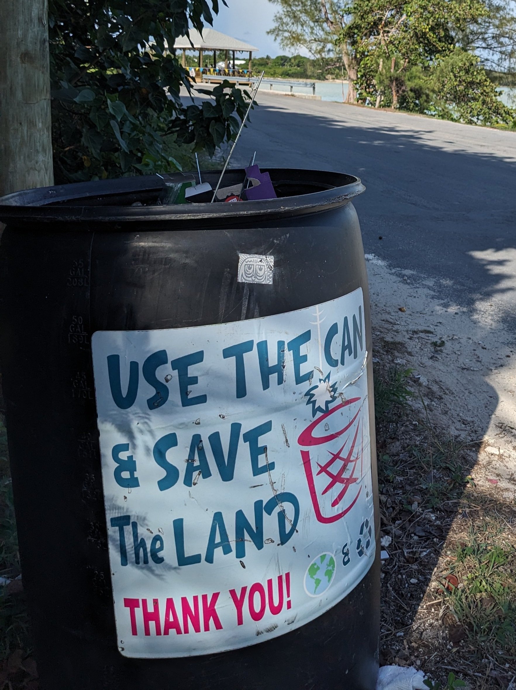 A large black trash can with a sign that reads 'Use the Can & Save the Land, Thank You!' attached to it; the sign also features a drawing of a trash can with a lid and a globe icon.