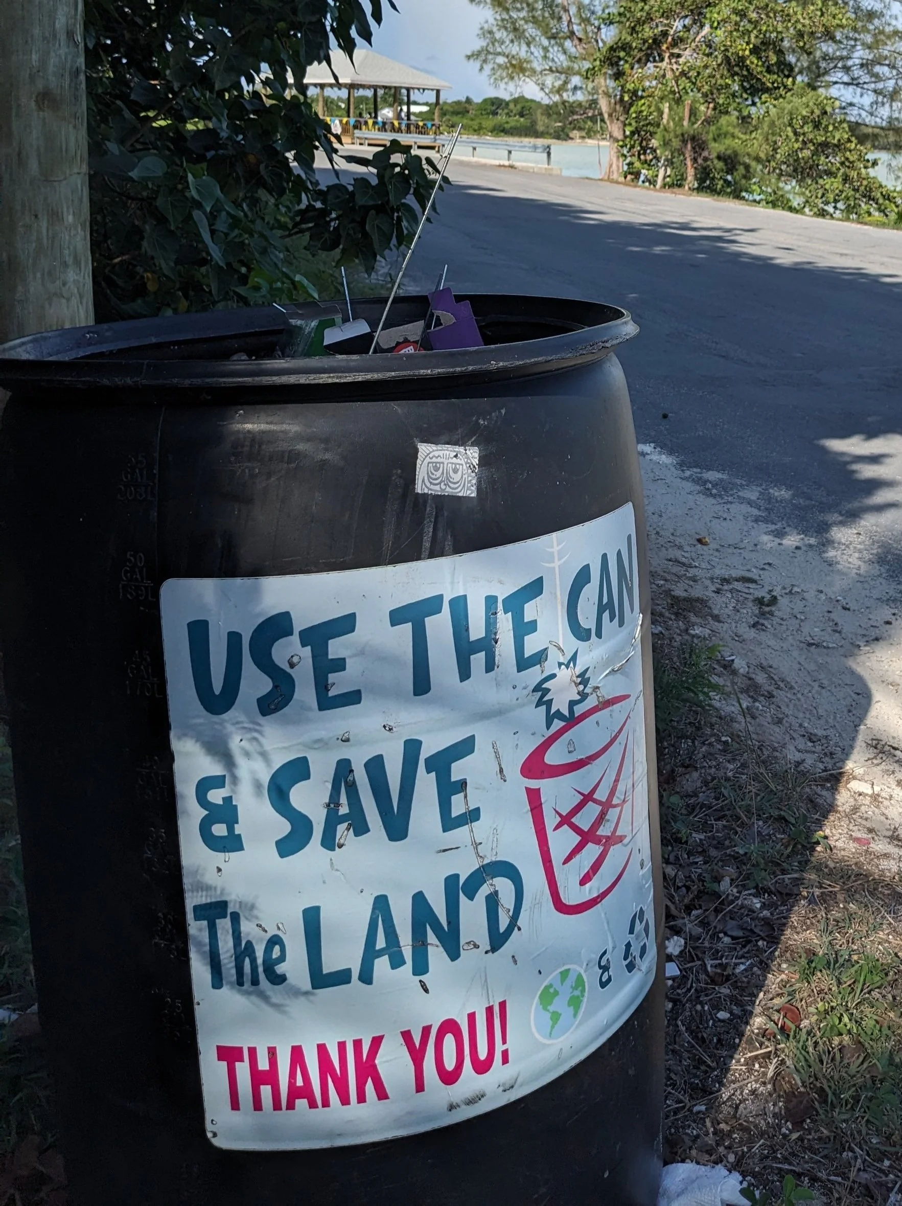 A large black trash can with a sign that reads 'Use the Can & Save the Land, Thank You!' attached to it; the sign also features a drawing of a trash can with a lid and a globe icon.