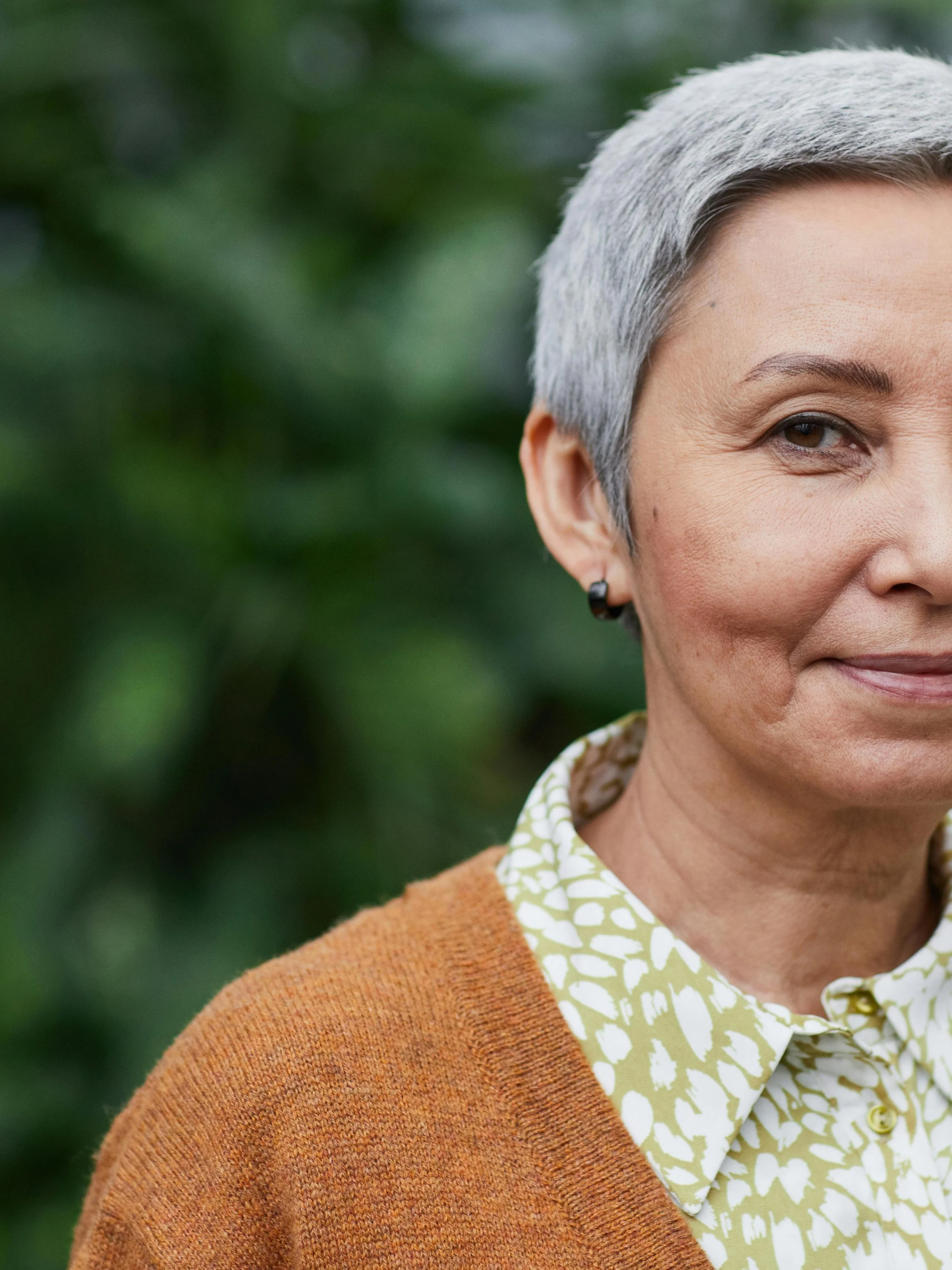Close-up of a mature woman with short gray hair, wearing a patterned collared shirt and an orange sweater, standing outdoors with greenery in the background.