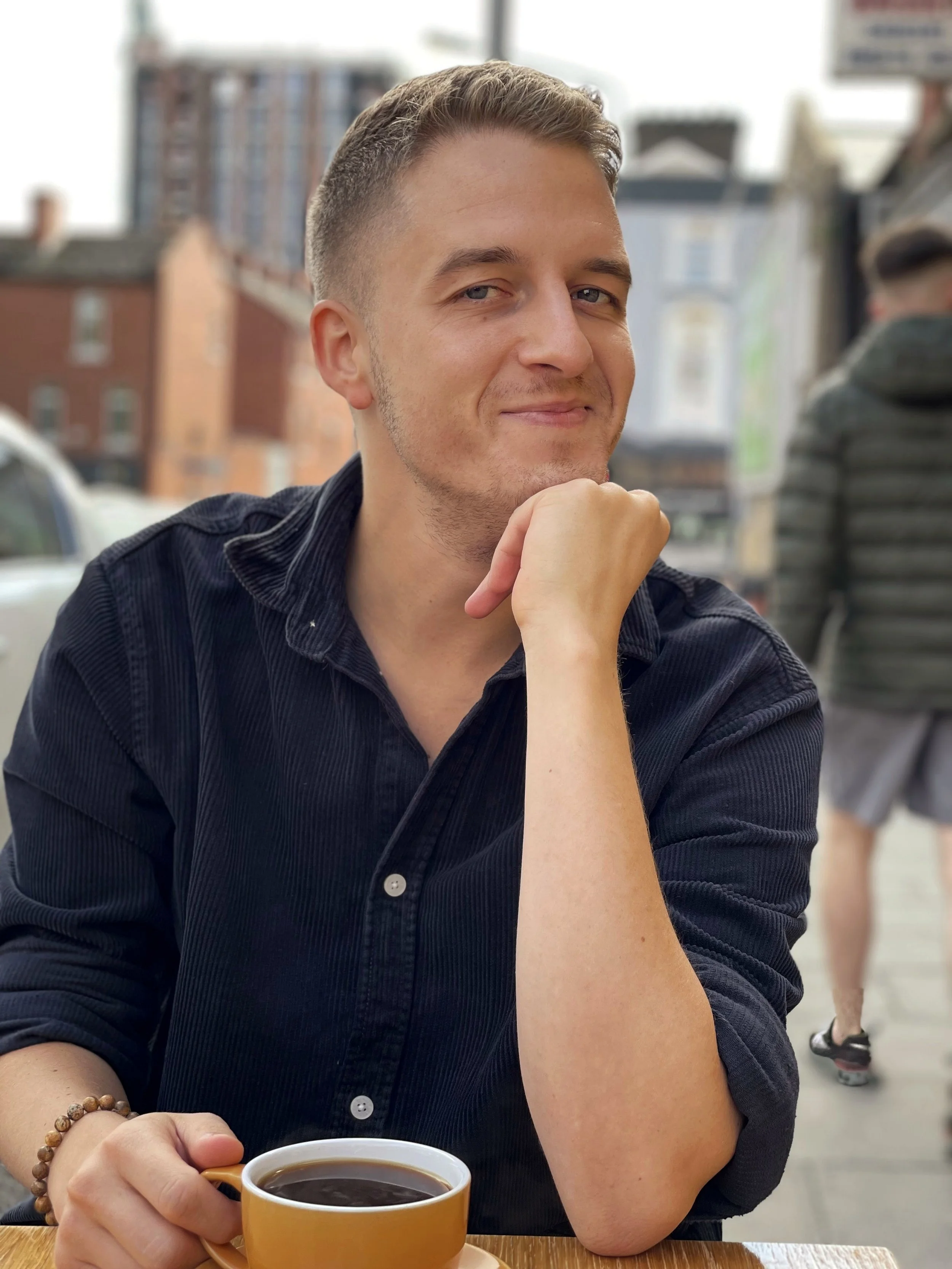 Man with short hair and light skin sitting at a table outdoors, holding a yellow coffee cup, smiling, with buildings and a person walking in the background.