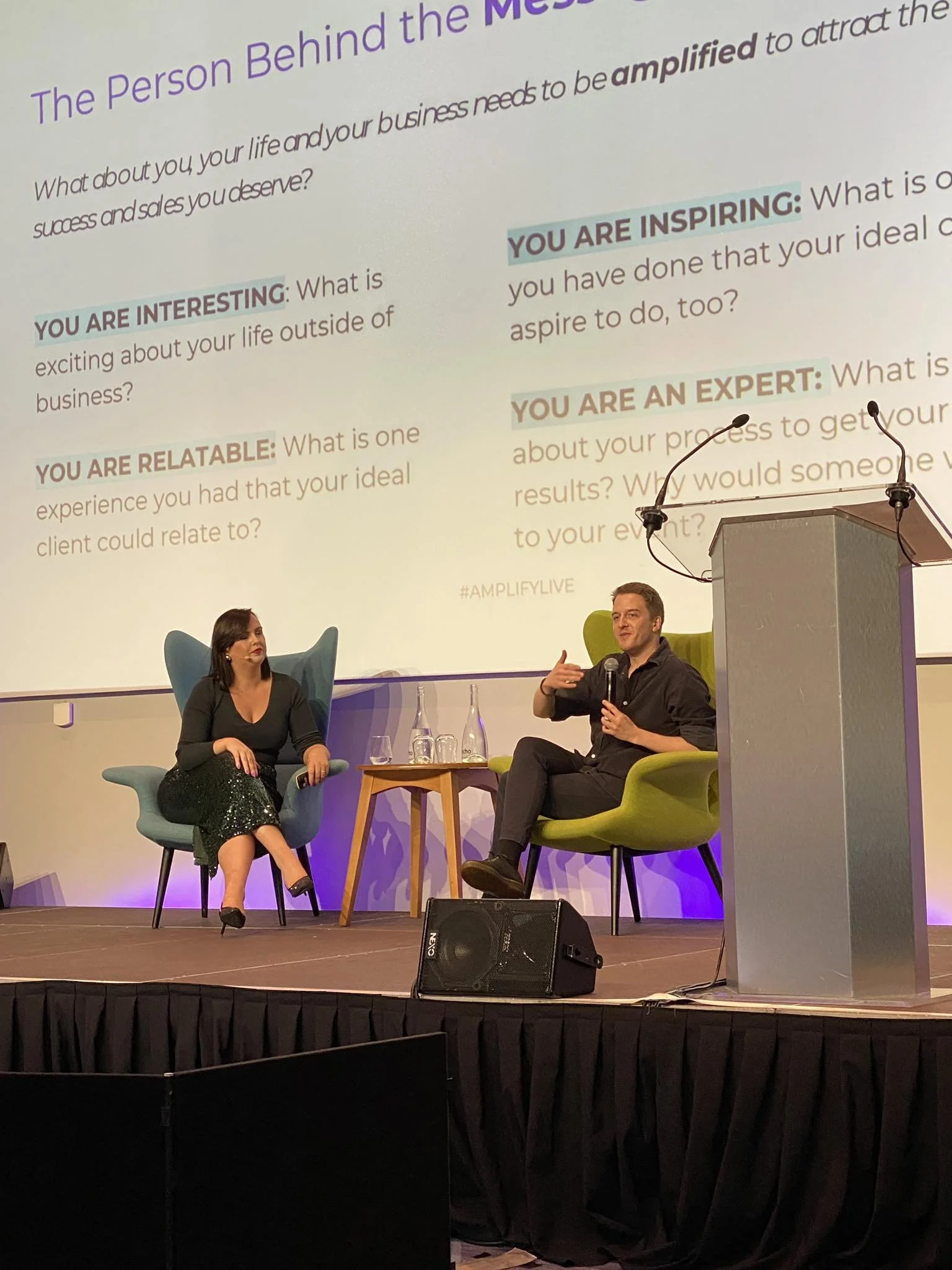 A man and a woman sit on stage in front of a large projected presentation slide. The man is speaking into a microphone, gesturing with his hand, and the woman is sitting with her legs crossed, listening. The stage has two chairs, a small table with water bottles and glasses, and a podium with microphones. The slide behind them contains questions about personal and professional growth, including prompts like "What is exciting about your life outside of business?" and "What is one experience you had that your ideal client could relate to?"