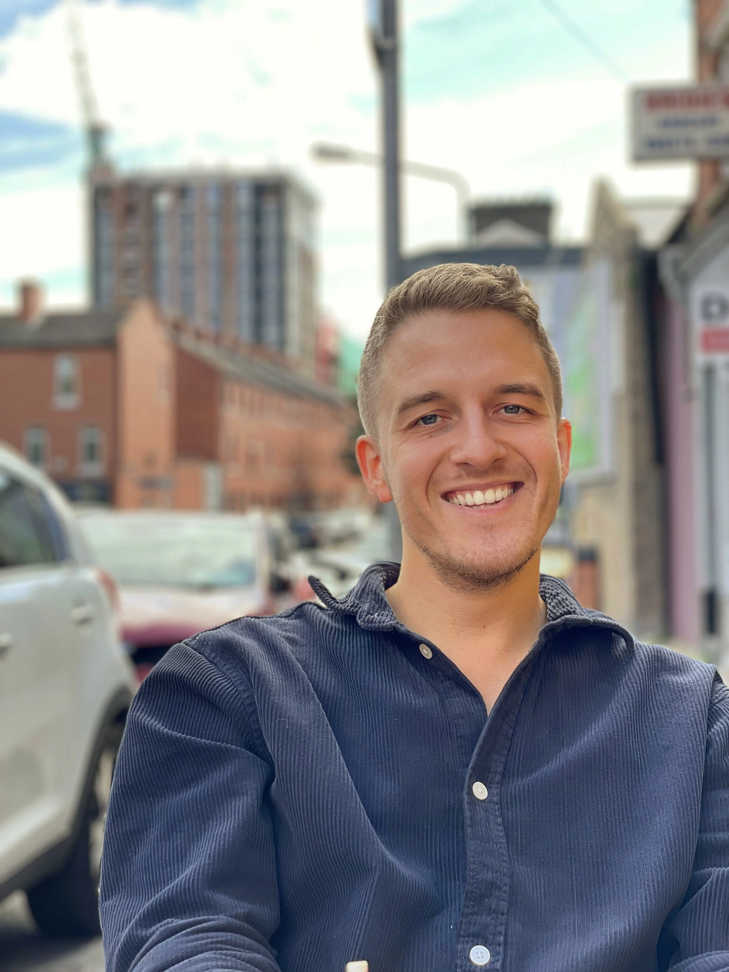 Young man smiling outdoors on a city street, wearing a dark blue button-up shirt.