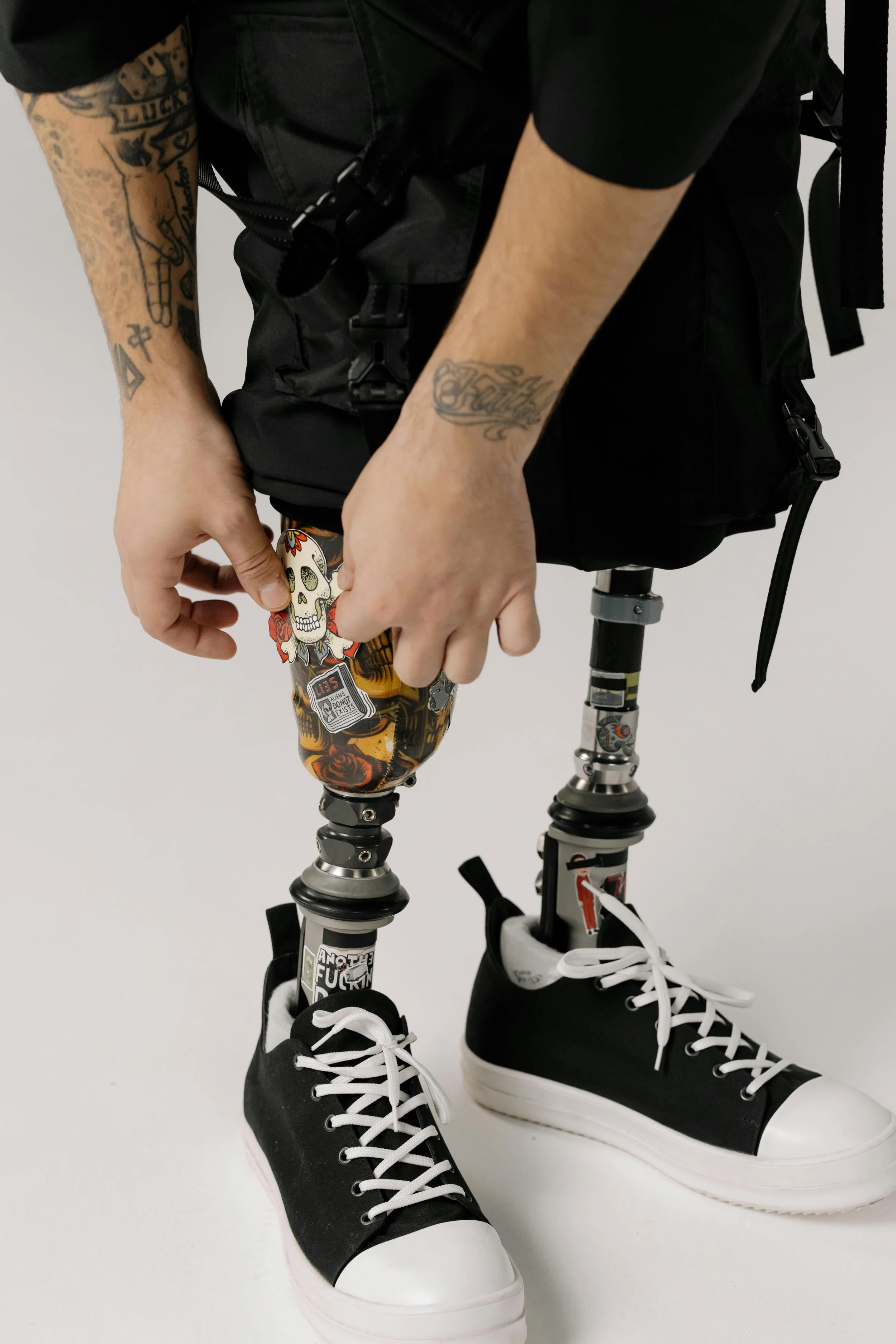 Person with tattoos on arms adjusting a prosthetic leg decorated with colorful stickers and wearing black and white sneakers.