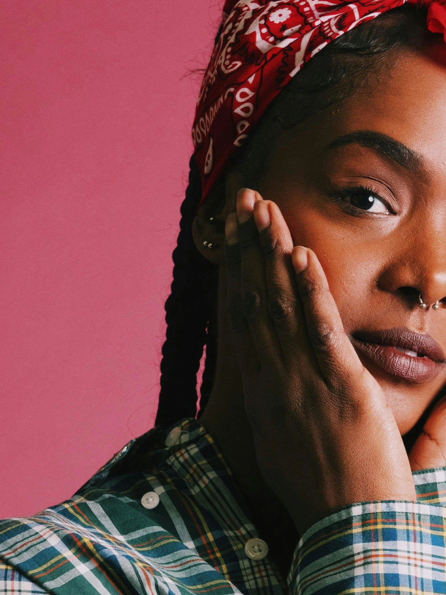 Close-up of a woman with dark skin touching her face, wearing a red bandana and a plaid shirt against a pink background.