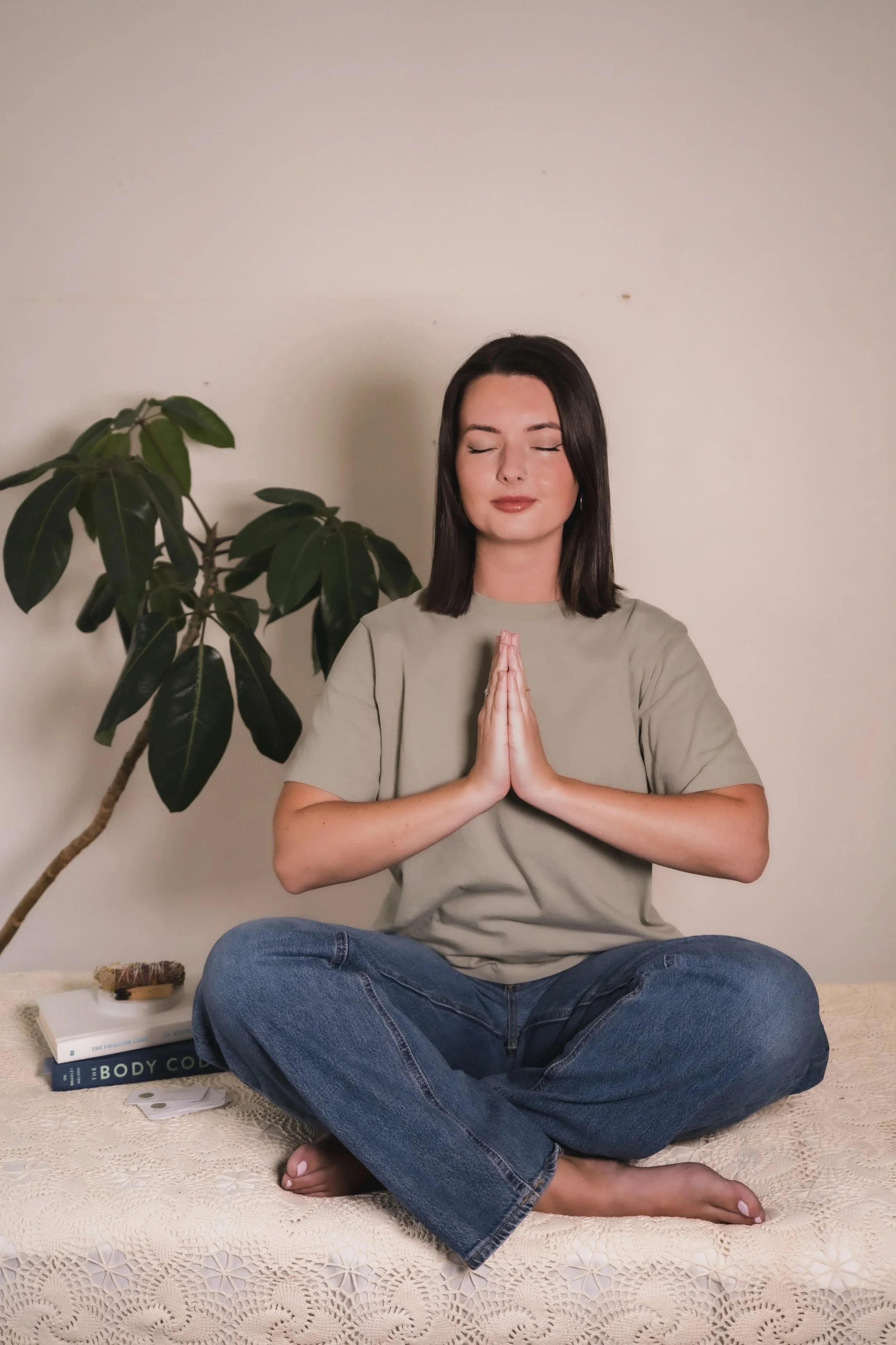 A woman with dark hair sitting cross-legged on a decorated table with a lace tablecloth, eyes closed, hands in a prayer position, next to a large leafy green plant. Books and a plate with a snack are nearby.