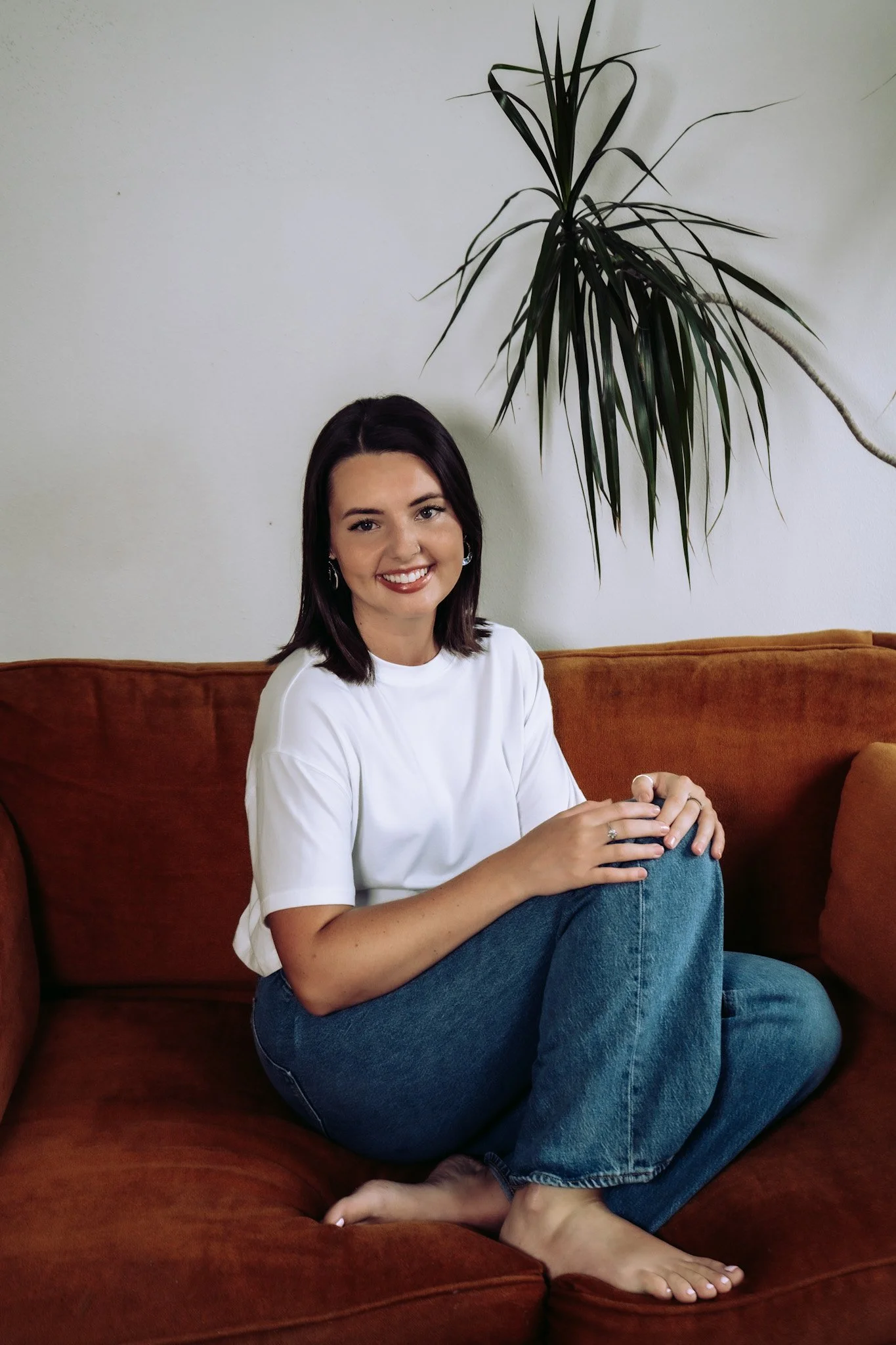 A young woman with dark hair sitting on a red velvet couch, smiling, wearing a white T-shirt and blue jeans, with a large green houseplant behind her against a plain white wall.