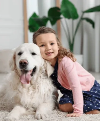 A young girl with curly hair in a pink cardigan and navy blue polka dot dress sitting beside a large white dog indoors, with green plants in the background.