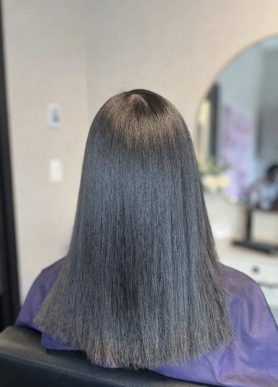 Back of a person's head showing long, straight, shiny, dark brown hair in a salon setting.