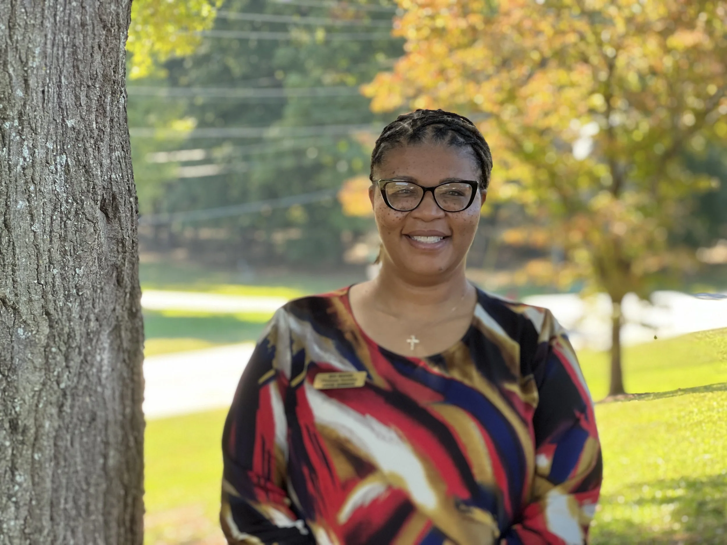 Woman standing outdoors next to a tree, wearing glasses and a maroon turtleneck sweater, smiling at the camera.